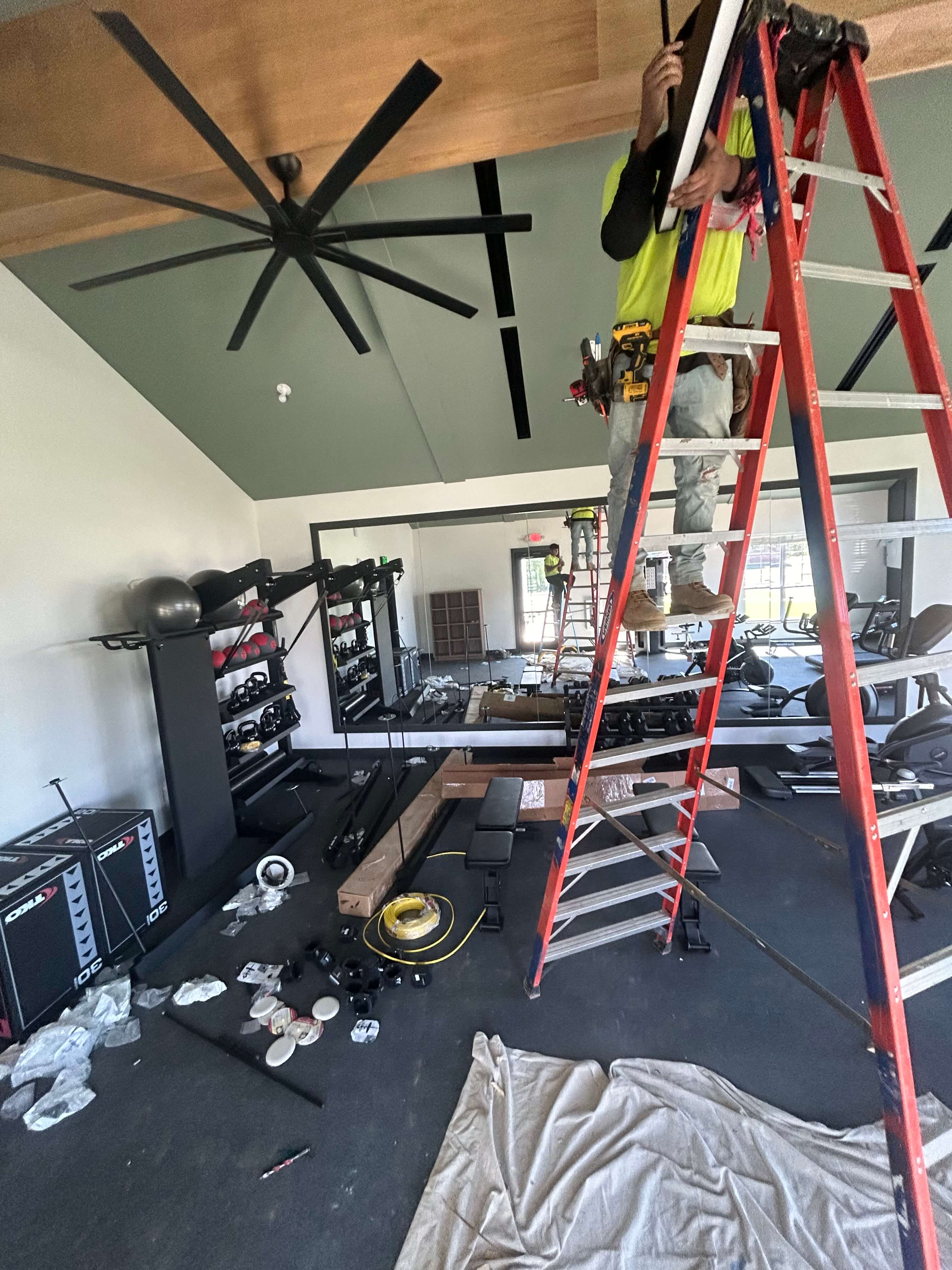 Electrician installing large ceiling fan in fitness center using ladder and tools